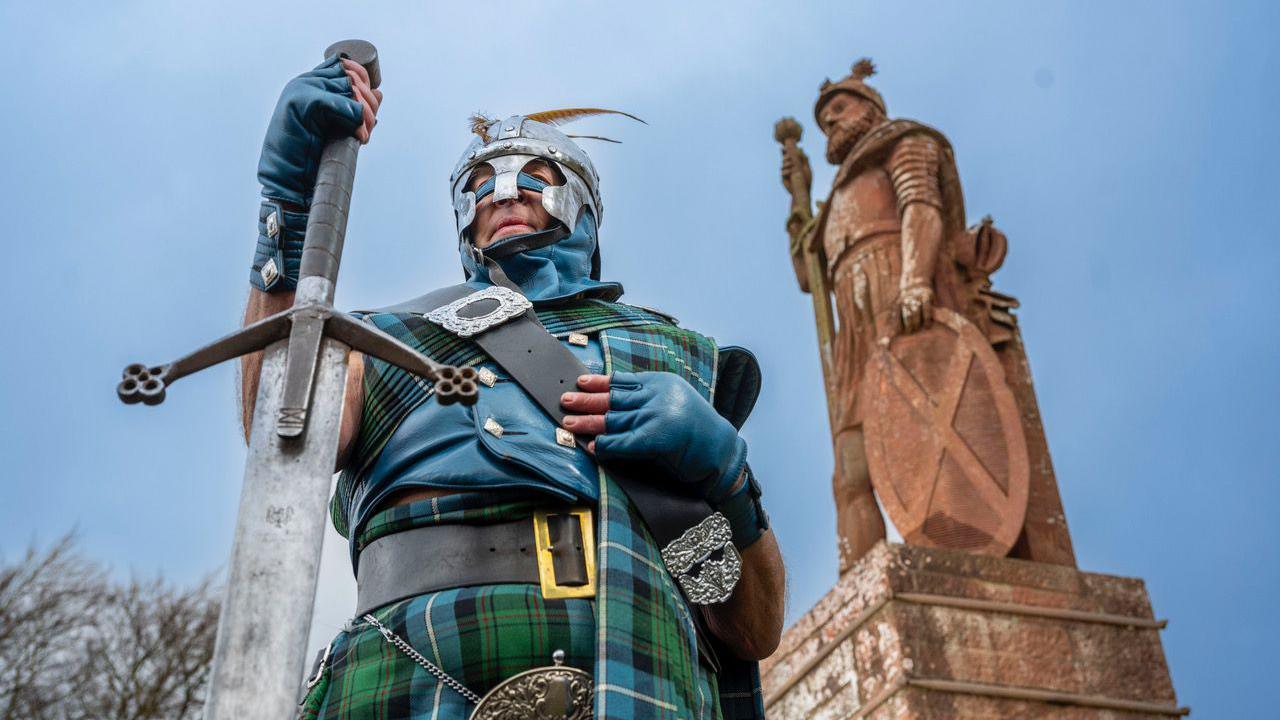 Jesse Rae - in full tartan dress with a helmet and large sword - stands in front of a sandstone statue of William Wallace. There is a blue sky behind him.