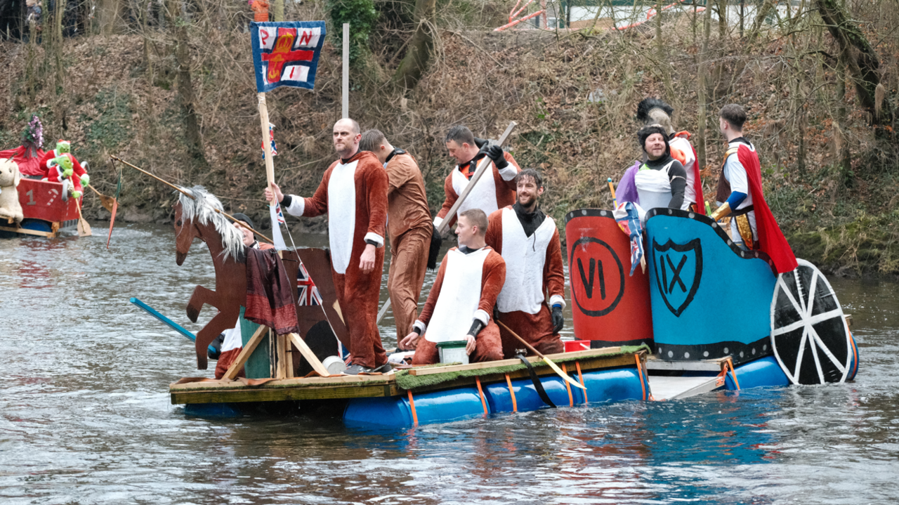 A medieval-themed custom-built raft with a number of people on it in fancy dress. The raft is floating along the River Derwent in Matlock.