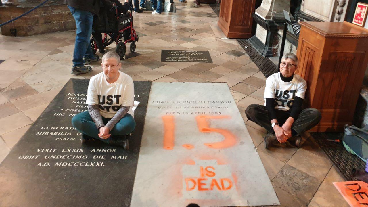 Two Just Stop Oil are seen sitting on the floor in Westminster Abbey at each side of Darwin's marble nave wearing white T-shirts with the group's name on it. "1.5 is dead" is painted on in large orange font.