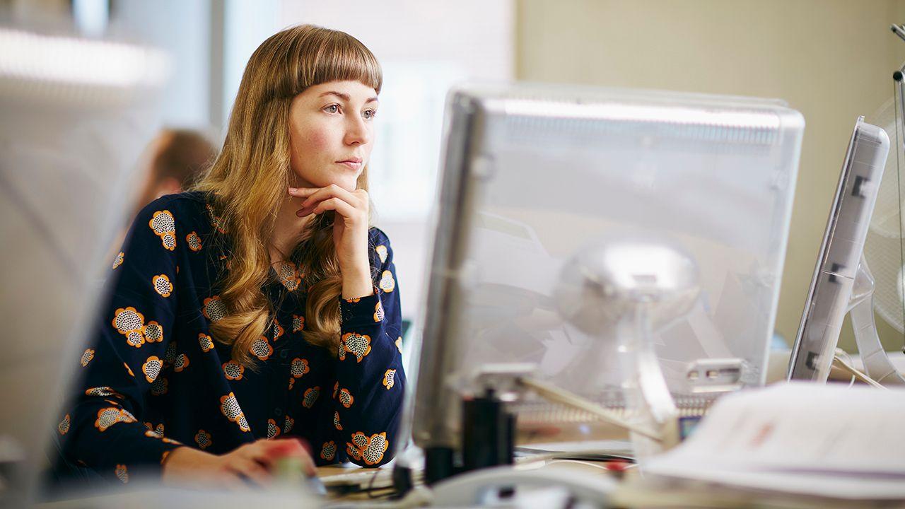 A woman looks at a computer screen in an office while using the mouse, she has blonde hair and looks pensive while wearing a blue and orange patterned shirt.