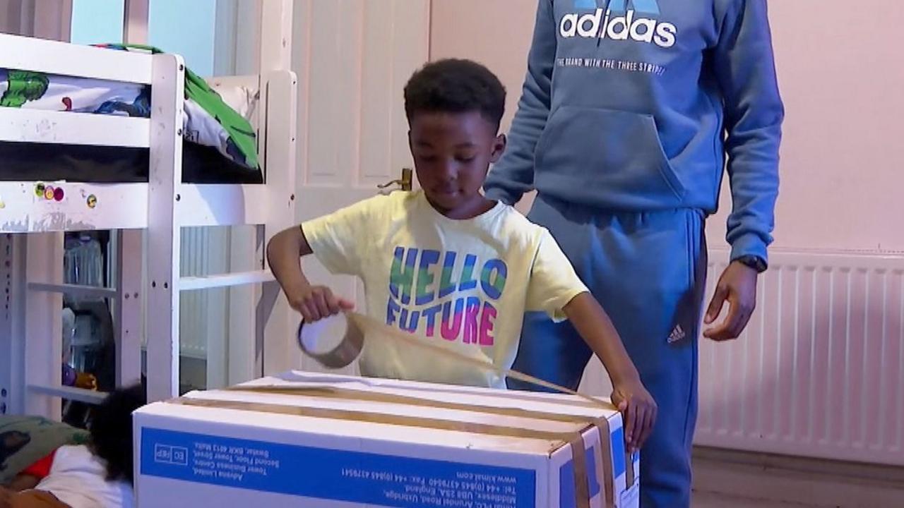 A child in a yellow shirt uses tape on a cardboard box while his father watches in a bedroom with bunk beds in the background.