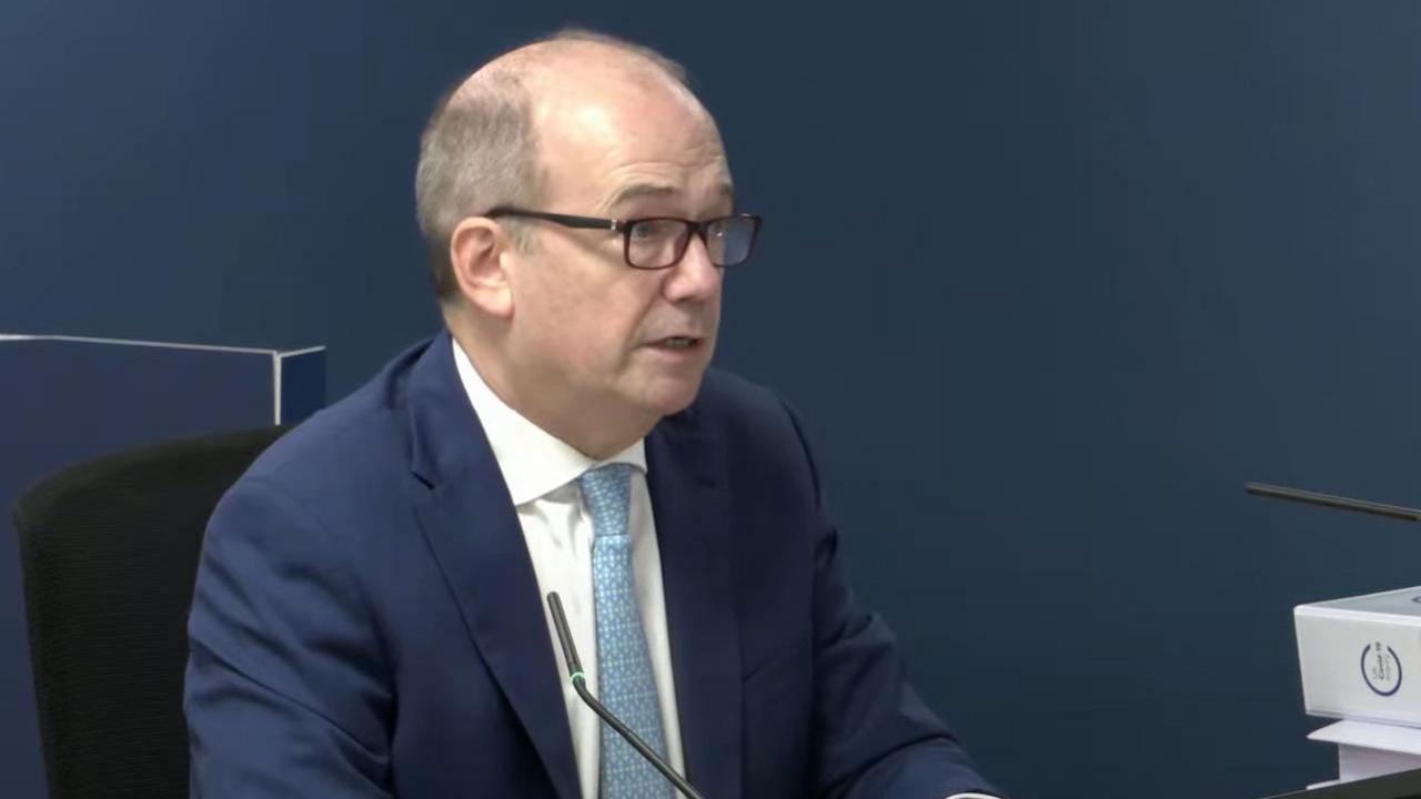 A screenshot of Dr Michael McBride giving evidence at the Covid Inquiry in London. He is wearing a navy suit jacket, a white collared shirt and a blue tie.