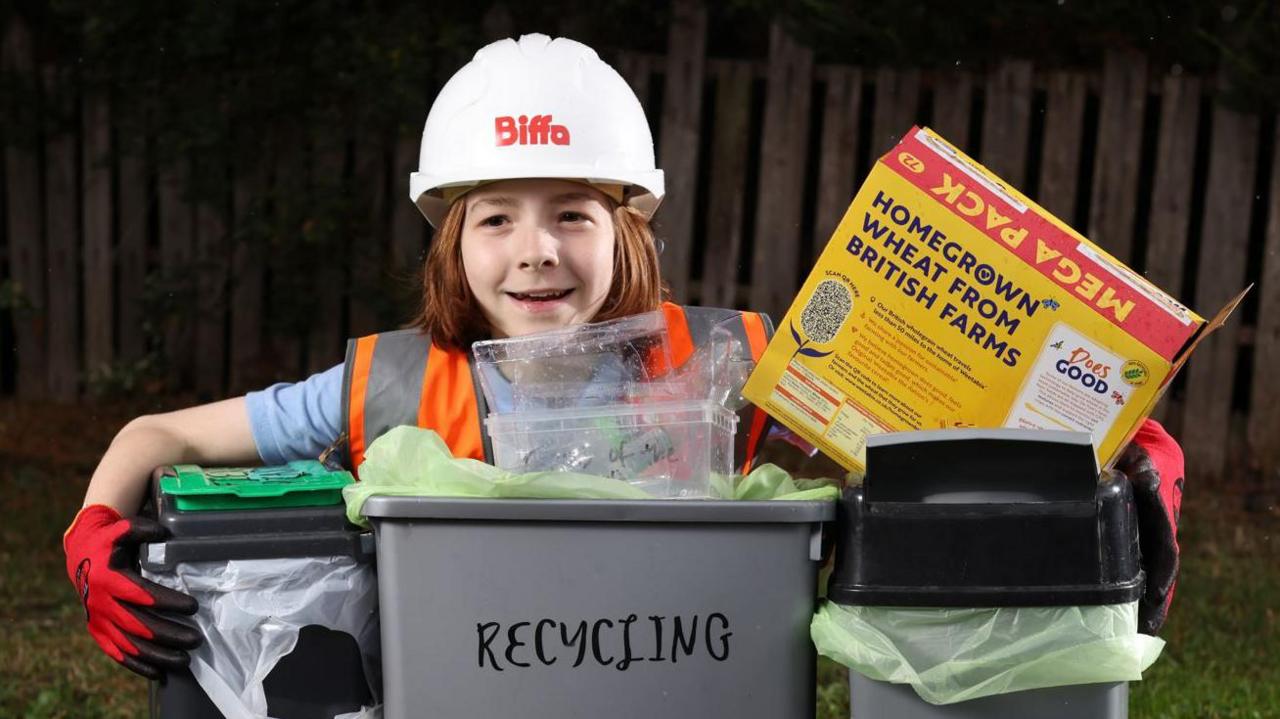 Tommy crouched behind three recycling bins holding a cardboard cereal box. He is wearing a high visibility orange bib, red gloves and a white hard hat with Biffa written on it in red writing