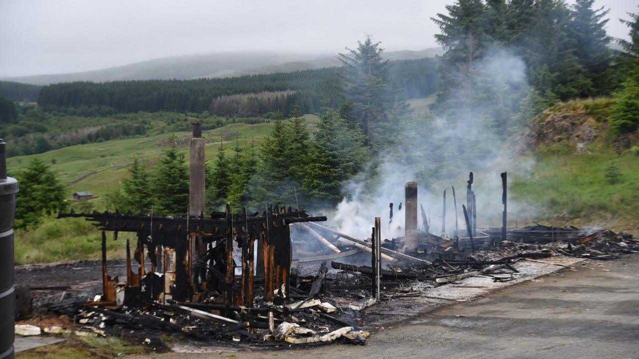 The smouldering remaids of a building stand on tarmac in the middle of a tree-lined country scene