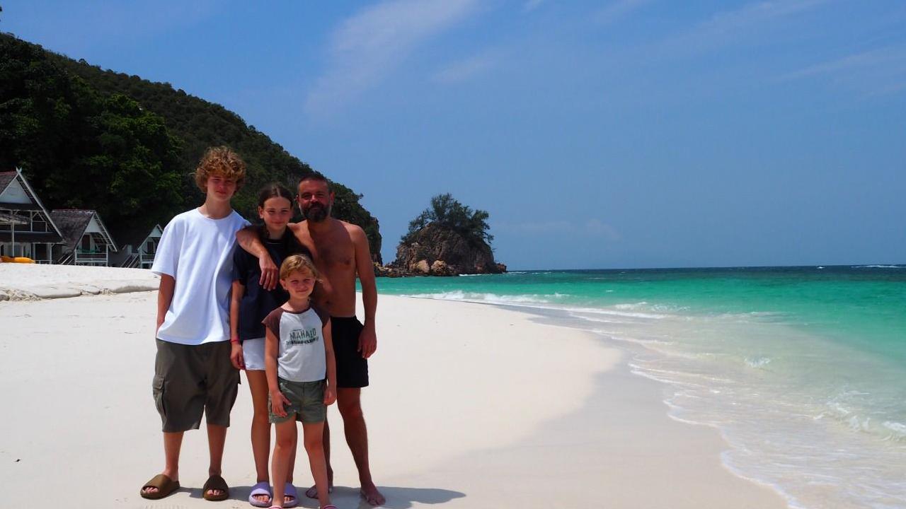 Geraint and his three children one a white sandy beach with huts in the background and the blue sea behind them.