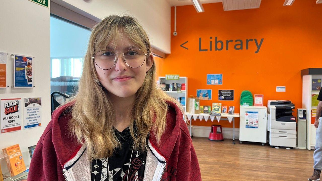 Sawyer, wearing round-rimmed glasses and a black and white V-neck sweater with a burgundy hoody with fleeced lining. A sign for the library is on the back wall behind her.