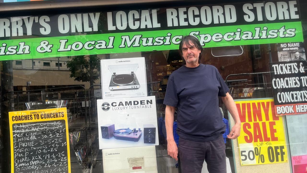 A man in a blue t-shirt, with dark long hair, stands in front of a shop window, with signs show discount record sales amd cpaches to concerts