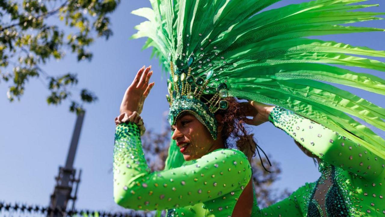 A woman in a green leotard and a green jewelled headdress holds her hand up to block the sun from her eyes.
