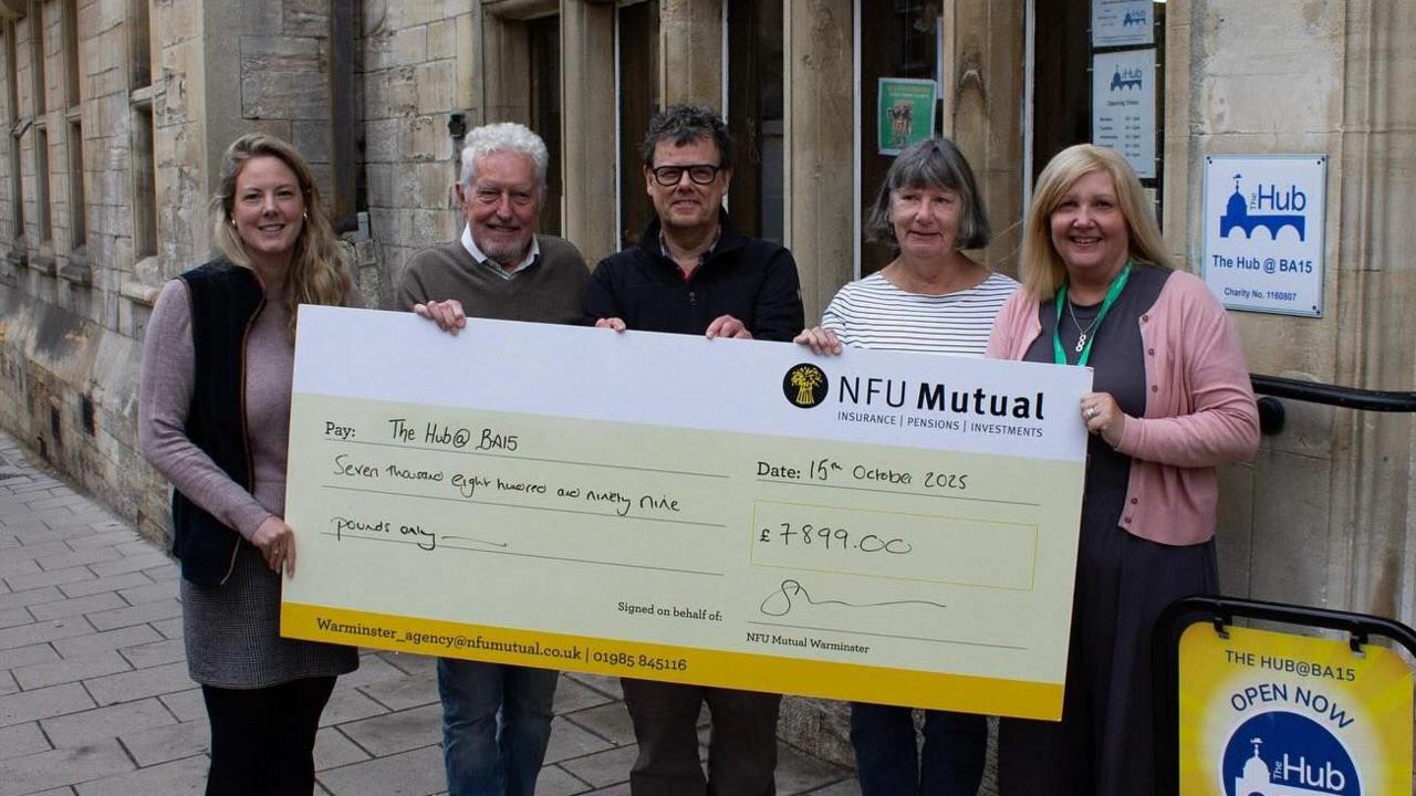 Five people smiling on a street and holding a big cheque which says 'NFU Mutual' and has the amount of £7899.00 written in the figure box.