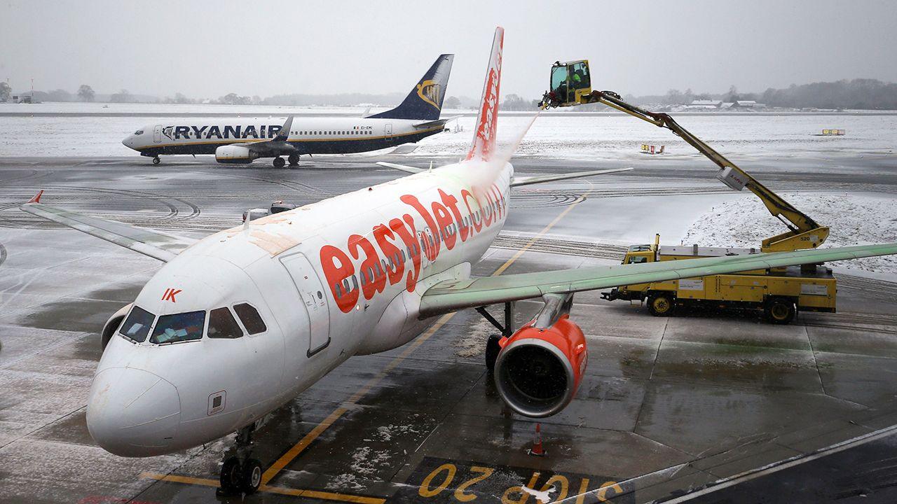 An Easyjet branded aeroplane is de-iced by an automated crane-style cart during cold weather, with a Ryanair branded aeroplane on the runway in the background, and with snow and ice all around, at Luton airport in 2017.