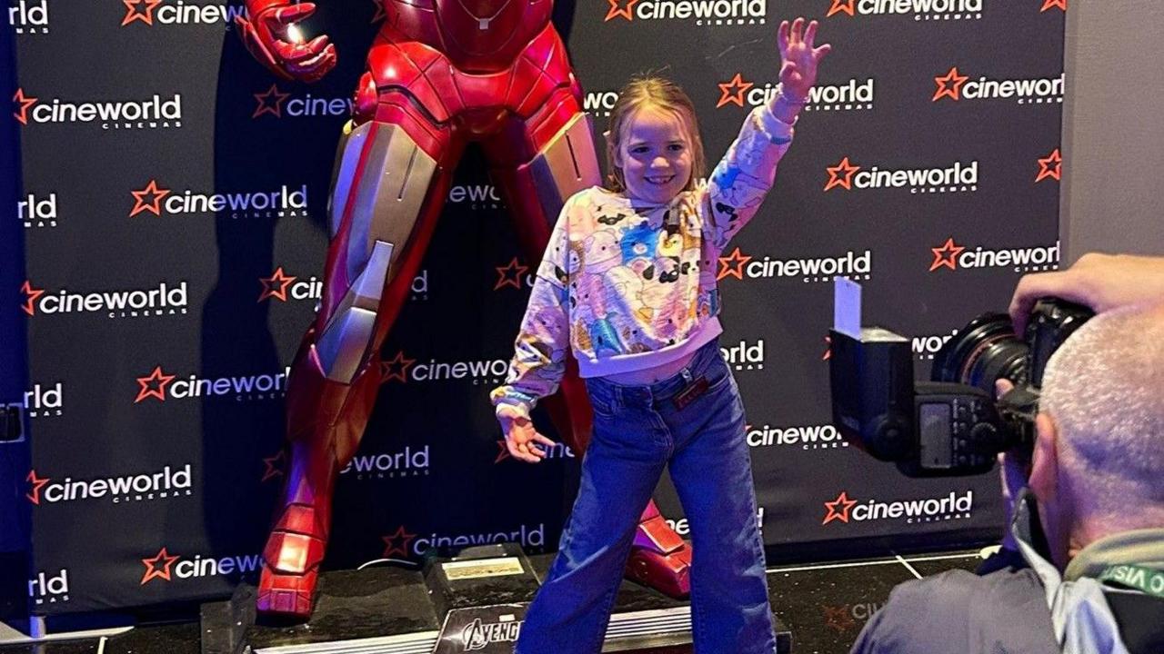 Ellie is stood with one hand almost on her hip and the other hand up in the air, she is smiling and posing for a camera in front of a superhero with a Cineworld banner behind her.