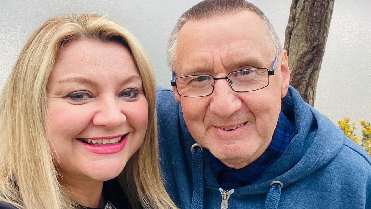 A woman with long blonde hair smiles and cuddles up to an older man with cropped grey hair and glasses