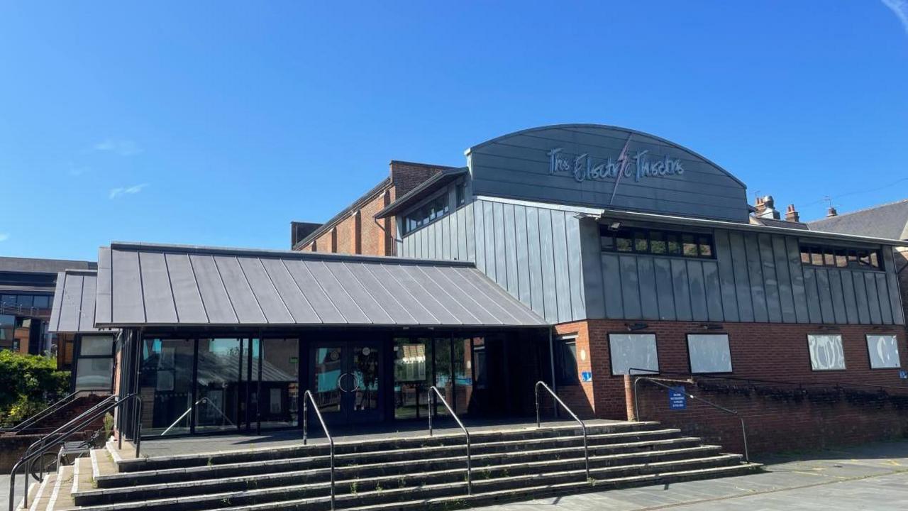 A large building made from bricks and grey panelling. It has a large sign reading The Electric Theatre and blank spaces that typically would advertise upcoming shows.