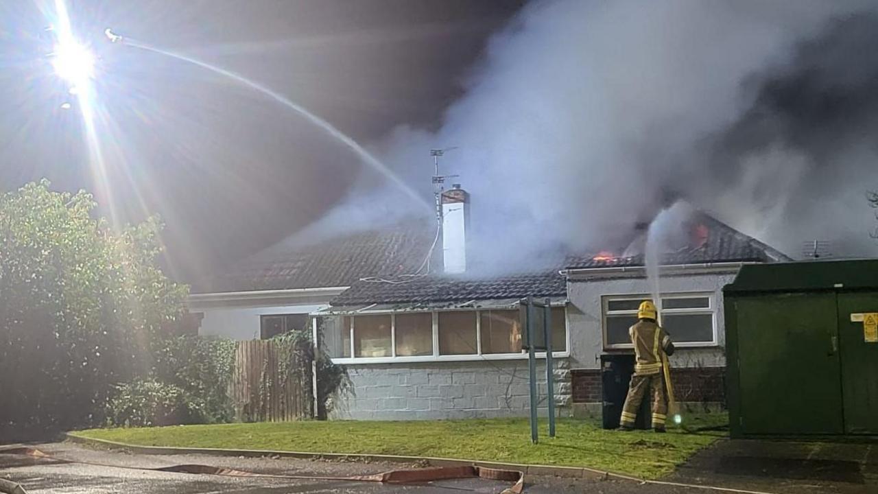 A firefighter wearing a yellow helmet points a hose reel jet towards flames coming out of the roof of a white bungalow. It is dark and the smoke billowing from the building appears white as it is lit by floodlights on a cherry picker above the bungalow.