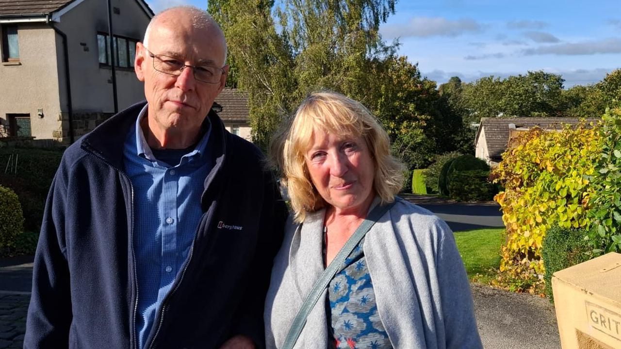 A man and woman look into the camera with neutral expressions on their faces. Behind them are a few houses surrounded by hedges and trees.