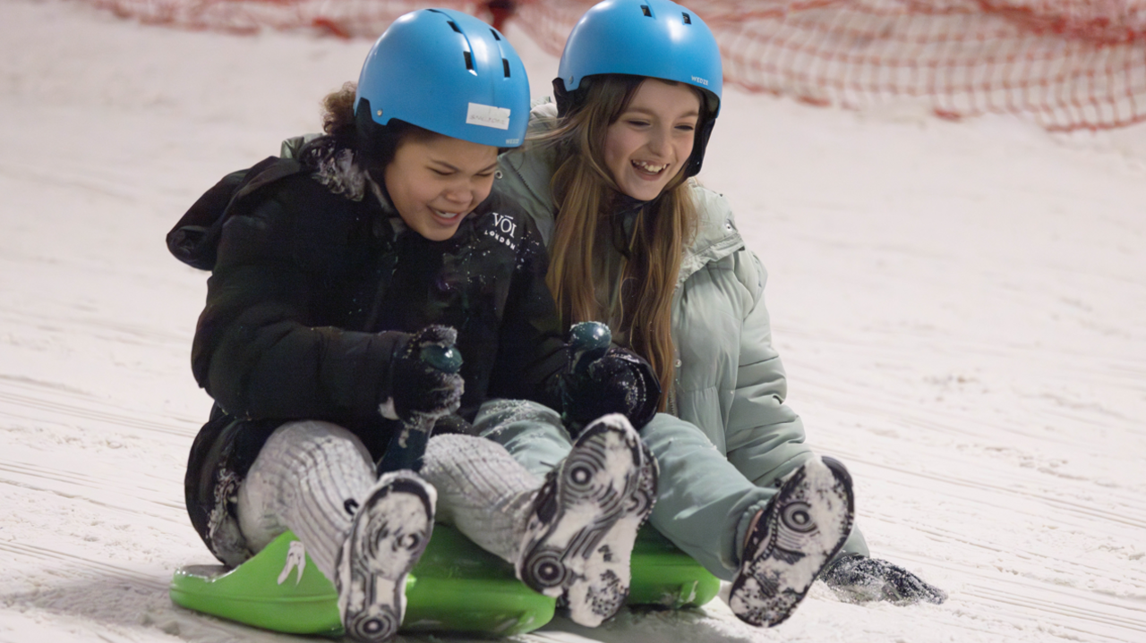 Two young girls are sat on green sledges and travelling down a snowy slope. Their feet are off the ground and covered in snow and both are laughing as they go. They wear blue helmets and warm winter coats.