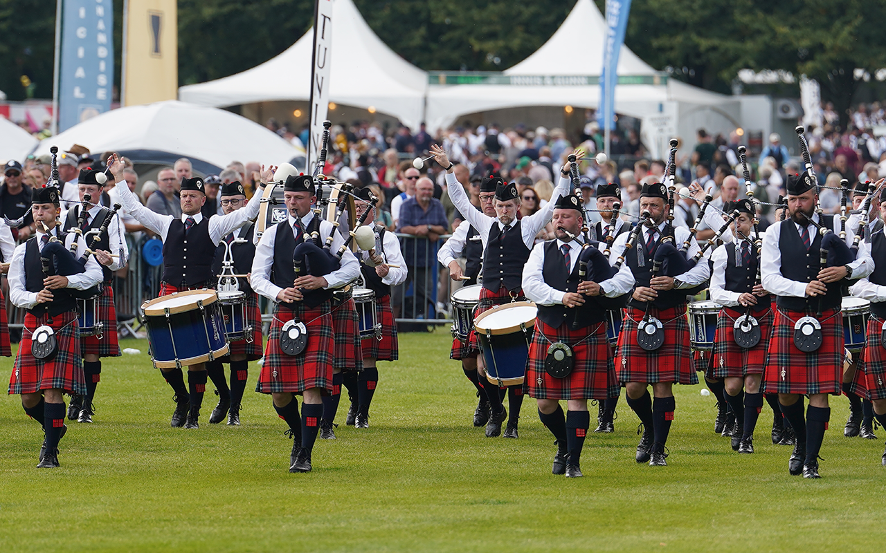 Pipe band in red kilts marching across a field with marquees and banners behind them