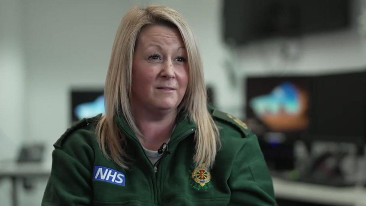 Gemma Robinson, a woman with mid-length blonde hair, wearing a green jacket with an NHS logo on it. The background is blurred but there are a couple of computer screens.