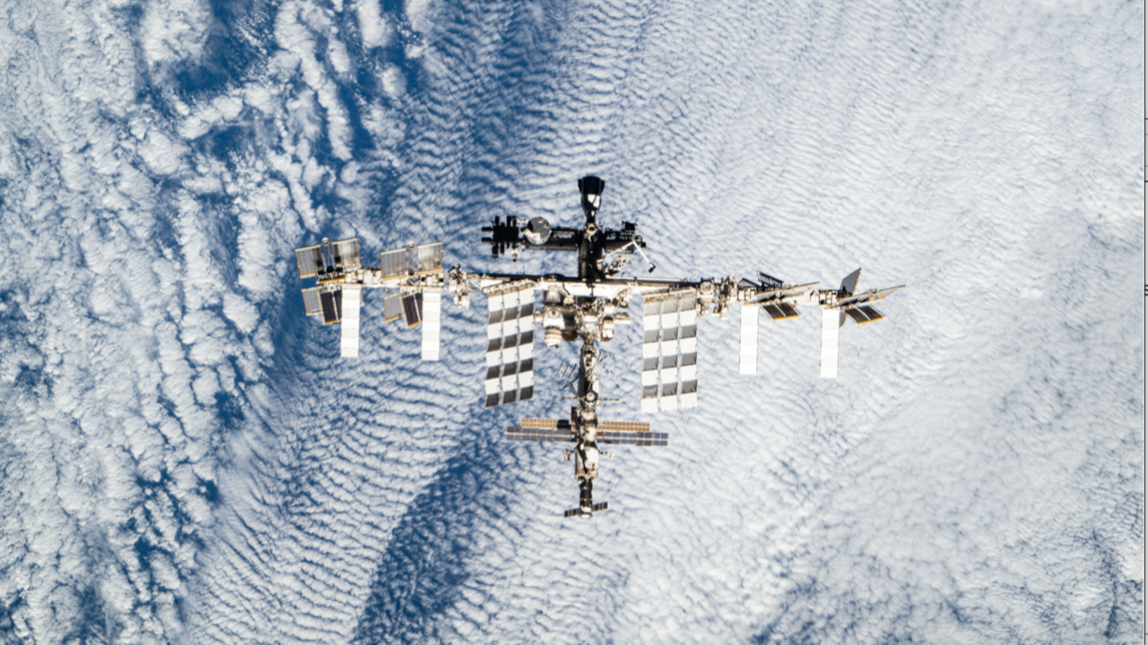 The picture shows the entire International Space Station seen from directly above, floating over a textured blanket of white cloud. The station’s long central truss runs horizontally across the frame, with large rectangular solar panels fanning out to top and bottom like wings. The modules and structures along the spine form a dense, metallic knot at the centre, while smaller radiators and panels extend at angles near the ends. Below, Earth’s clouds appear as rippled, swirling patterns in pale blue and white, giving a strong sense of height and the station’s fragile isolation in orbit.