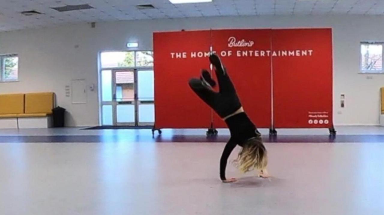 A woman halfway through a backflip. She is dressed in black, her back facing the camera. Behind her is a red banner with white text reading: "Butlin's, the home of entertainment".