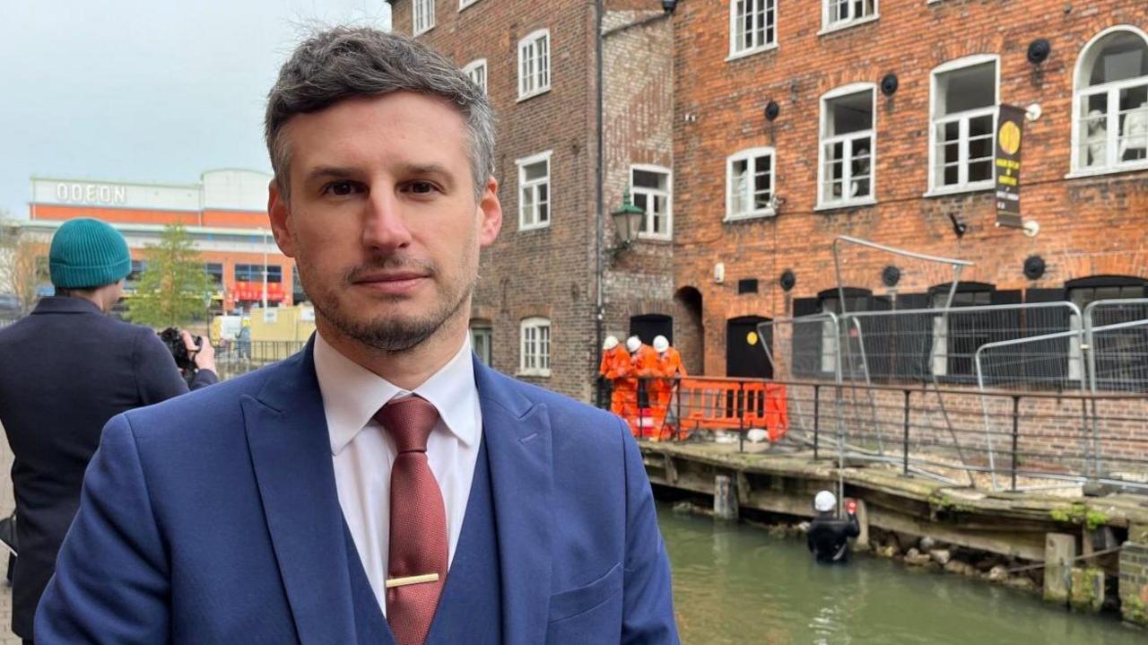 A man in a smart blue suit and red tie looks into the camera as he has his photo taken by a waterway. Divers and construction workers can be seen in the background.