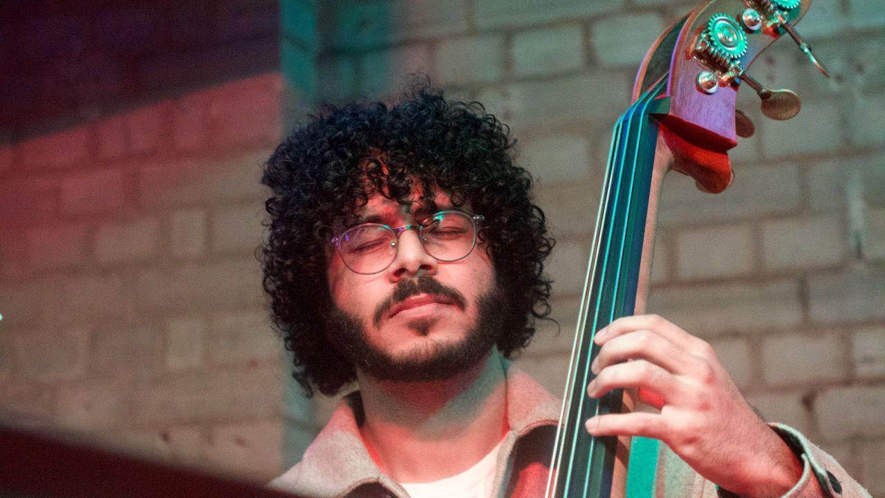 Shivraj has short brown curly hair, a beard and is wearing glasses. He is closing his eyes and playing the double bass.