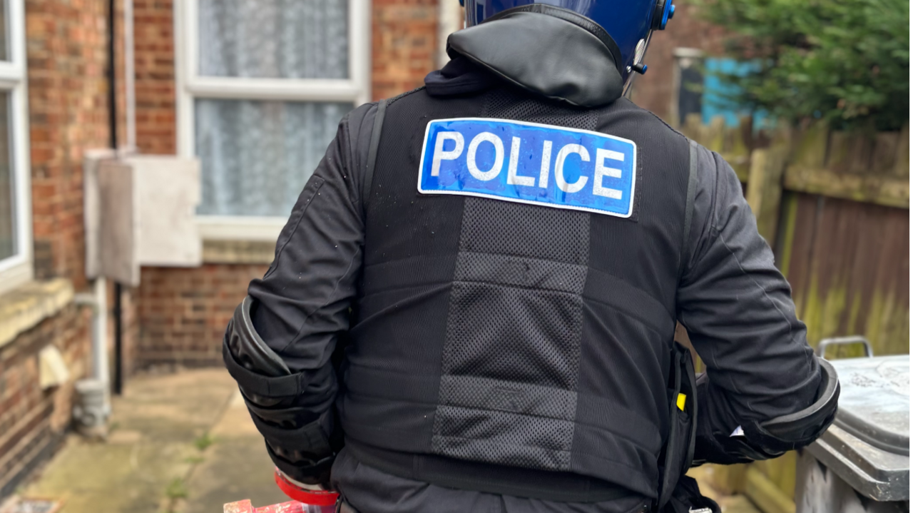 A close-up of the back of a uniformed police officer. He is wearing a blue helmet and a black uniform and is walking down a path at the side of a house. On the back of his jacket a blue badge says "POLICE". 