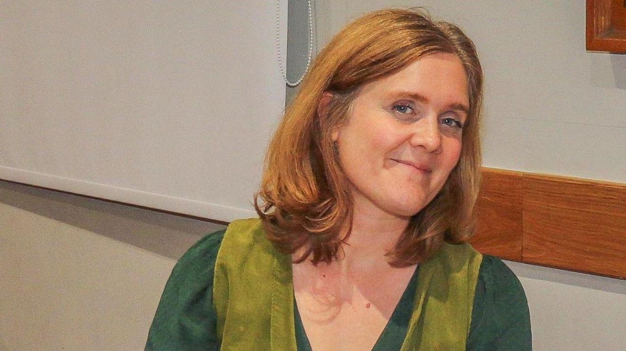 Holly Bembridge wearing two different shades of green, smiling. She is sitting by a wall, facing the camera, but with her face slightly tilted to one side.