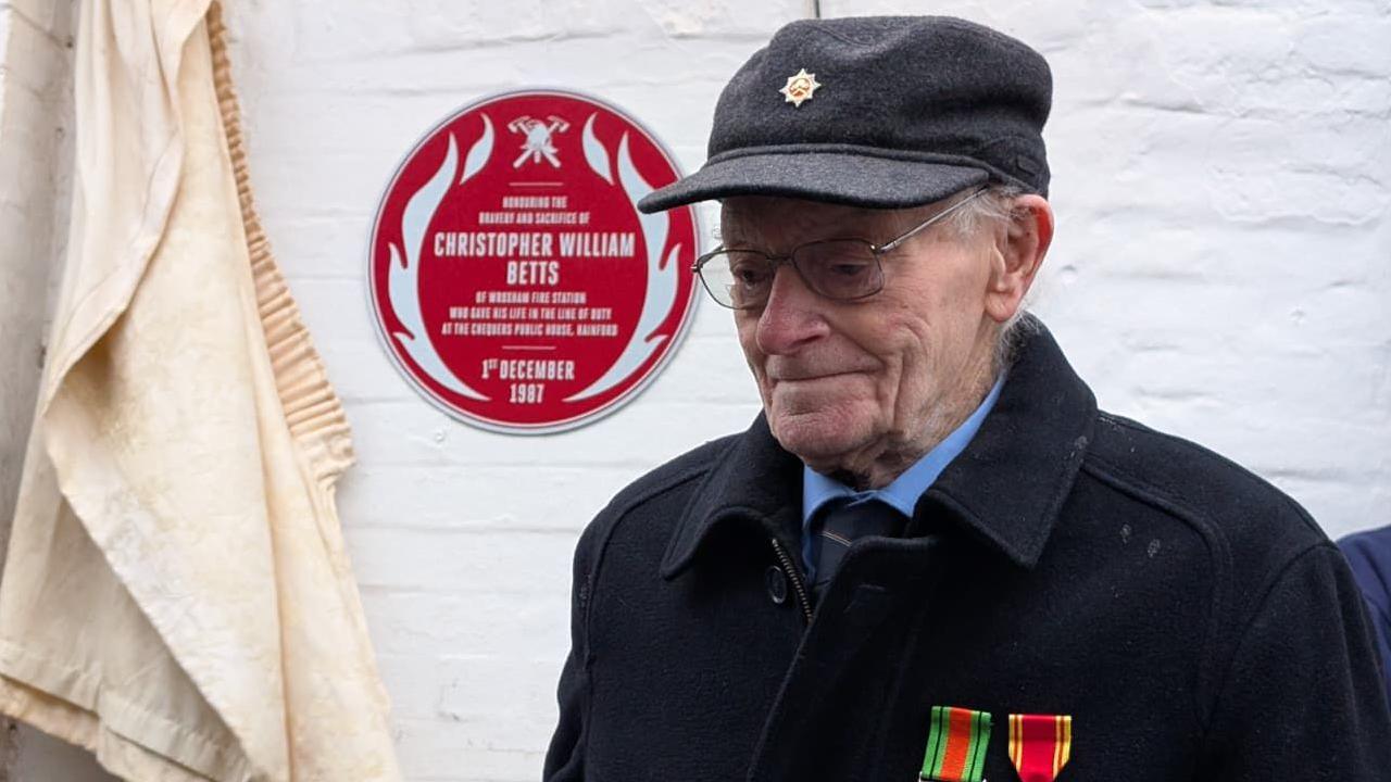 William Betts, an elderly man with glasses who is wearing a grey cap and a dark coat with two medals pinned to it, stands in front of a white brick wall. Over his shoulder, on the wall, is the red plaque paying tribute to his son, Christopher. The red plaque has white writing on it, which gives Christopher Betts' name and says when he died.