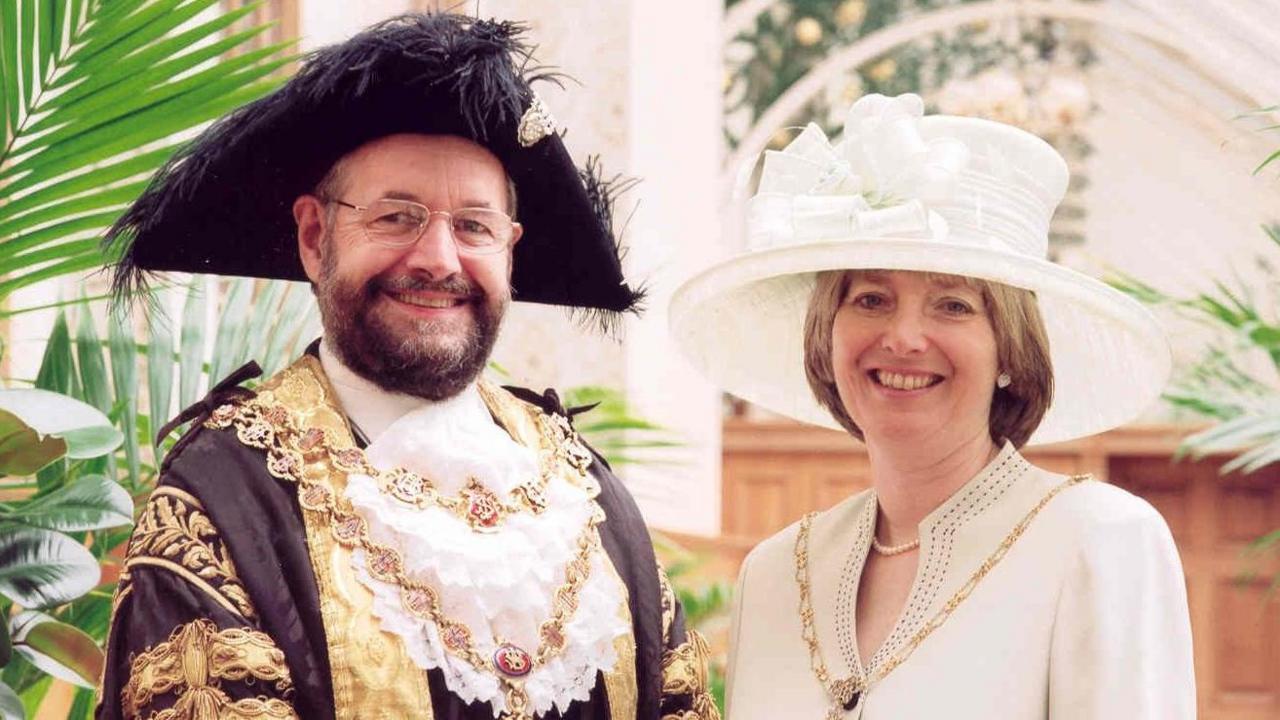 A man in lord mayor's attire - a black and gold outfit with a large black hat and gold chains around his neck - stands next to a woman wearing a white outfit, a large white hat and a smaller gold chain.