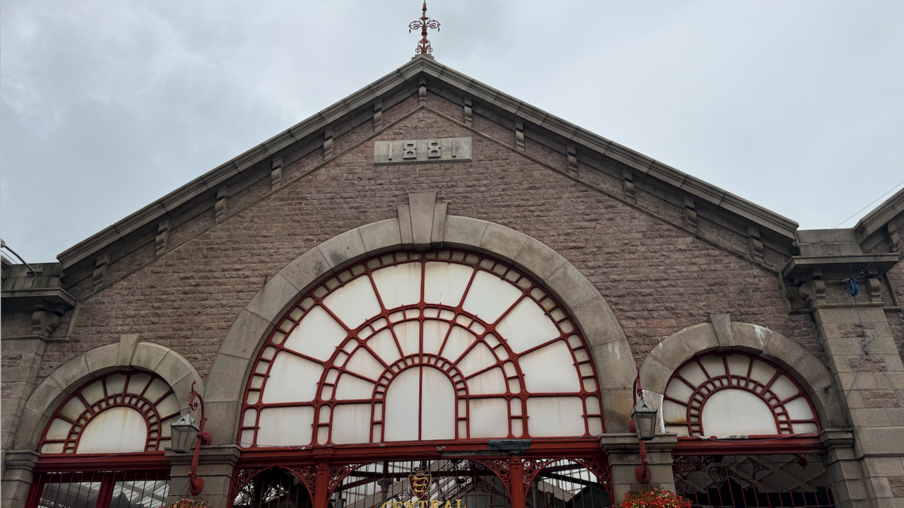 St Helier Central Market from the outside looking upwards, brown bricks with three semi circles, one big one in the middle accompanied by two smaller ones, all with red detailing, at the point of the building a sign saying 1881, on the point of the roof is a spire, grey skies behind