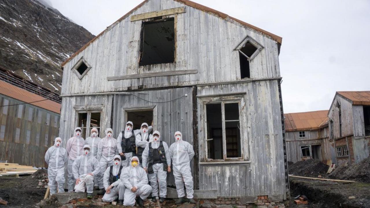 A group of eleven people in white hooded hazmat suits stand in front of an old wooden building
