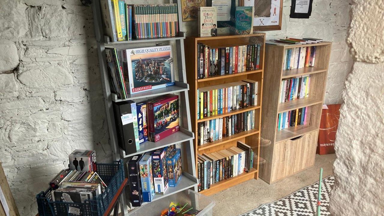 Three bookshelves. The one on the left is full of games and puzzles. The two to the right are filled with books. There is a crate of books to the left of the bookshelves. There is a black and white rug on the floor. 