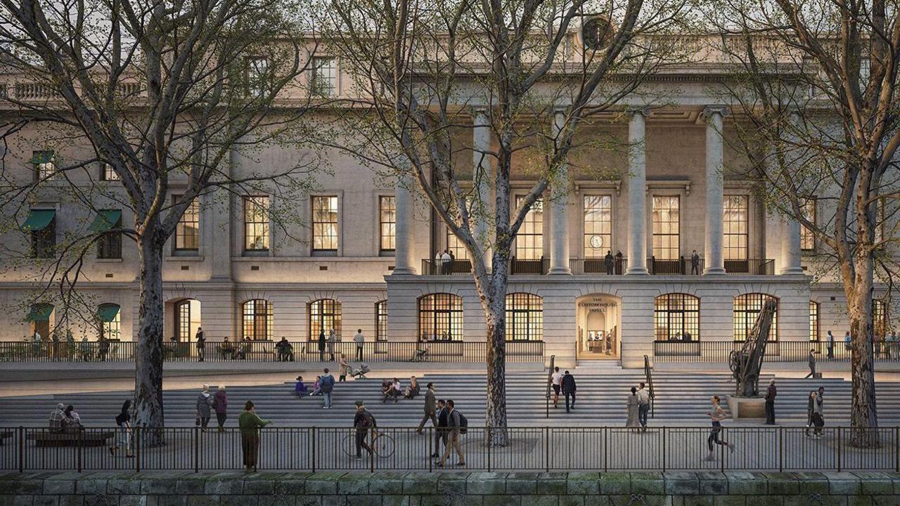 An artist's impression of the Custom House hotel redevelopment from the outside, showing the front facade of the building and public space along the river. The windows are lit up, and the pillars and front entrance of the building are visible. There are trees lining the public riverside walkway, and people are walking, cycling, running and sitting on the riverside.