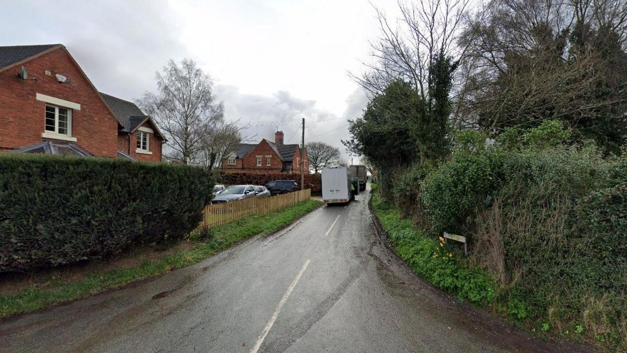 A generic image of a road, with bushes and trees to the left and right. There are buildings and vehicles parked to the left.