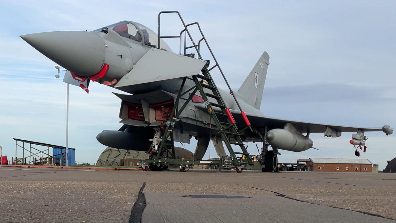 A Typhoon fighter jet stands on the tarmac at RAF Coningsby. Maintenance steps are standing next to the the grey coloured plane. A green aircraft hanger is visible in the background.