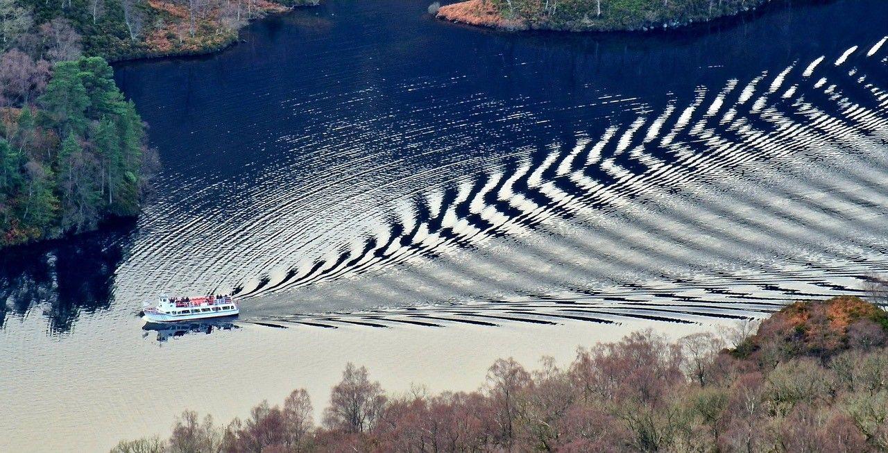 Boat going across a loch, leaving a ripple effect trailing behind it. 