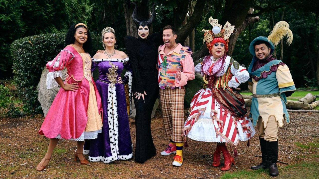 Six people in costume, with some in dresses, are standing in a line facing the camera. Trees and bushes are behind them.