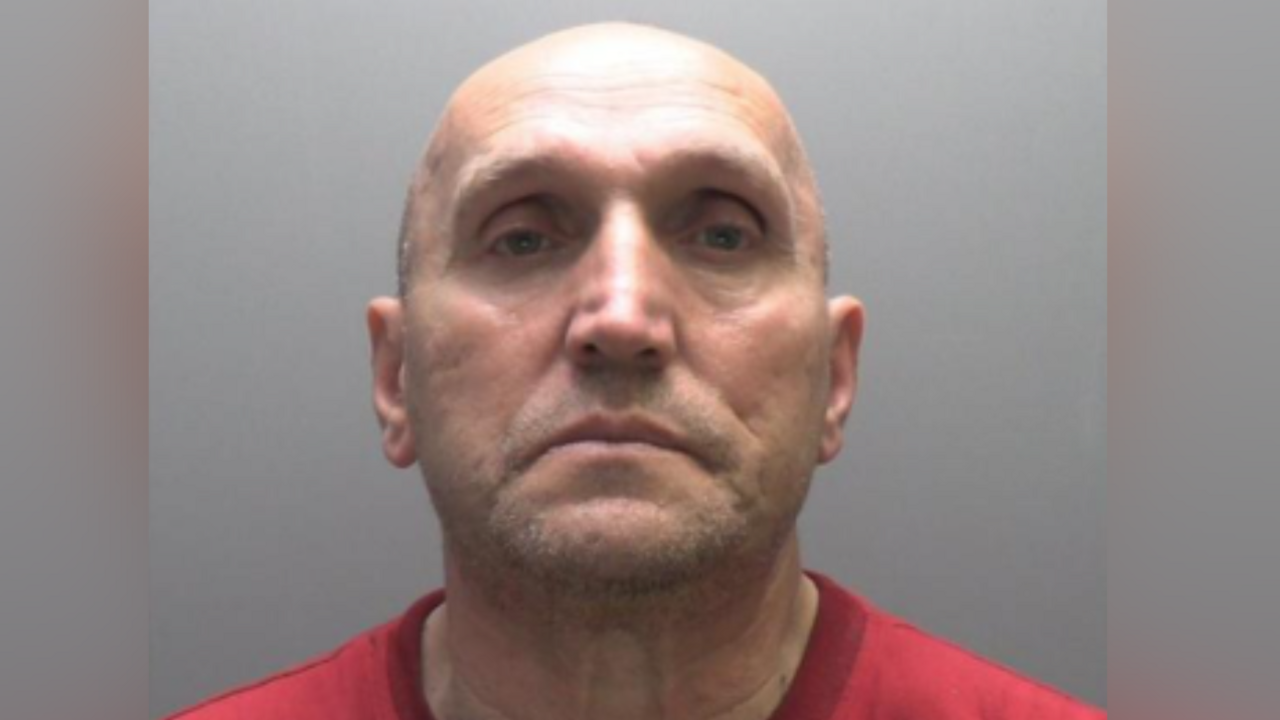 A mugshot of Iuliu Kubola, a bald man wearing a red T-shirt, looking at the camera with a frown. The photo is taken against a grey background.