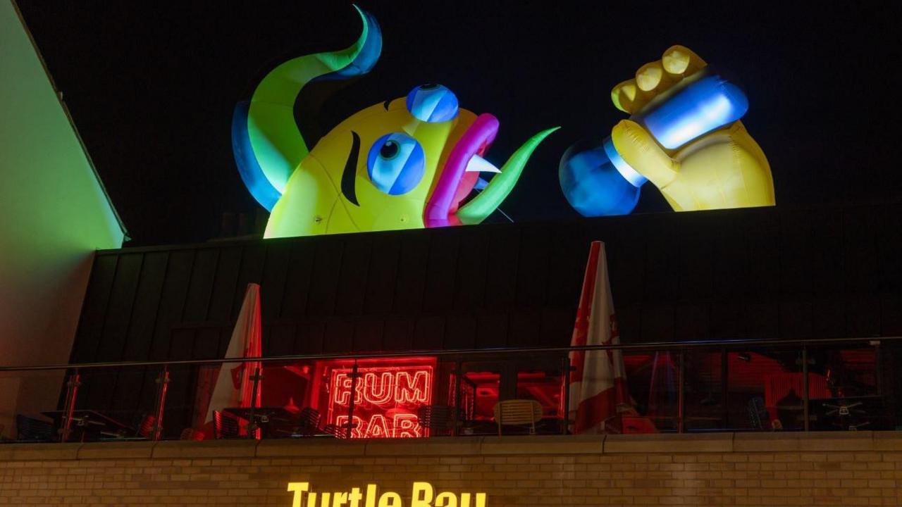 An outdoor bar with neon lights, tables, chairs and closed umbrellas. Behind is a large yellow monster with red lips and blue and green hair. The monster is holding a blue microphone to its mouth.