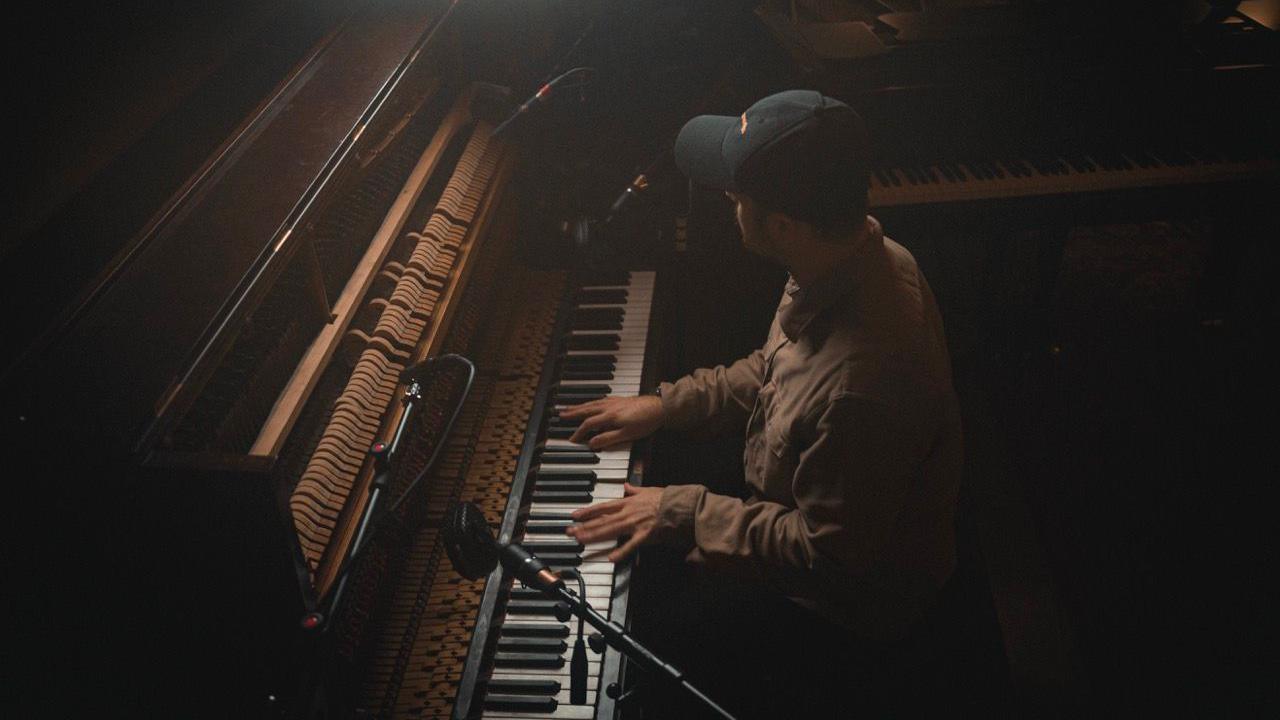 A man is wearing a black cap and a brown shirt. He is playing a piano in a darkly lit room.
