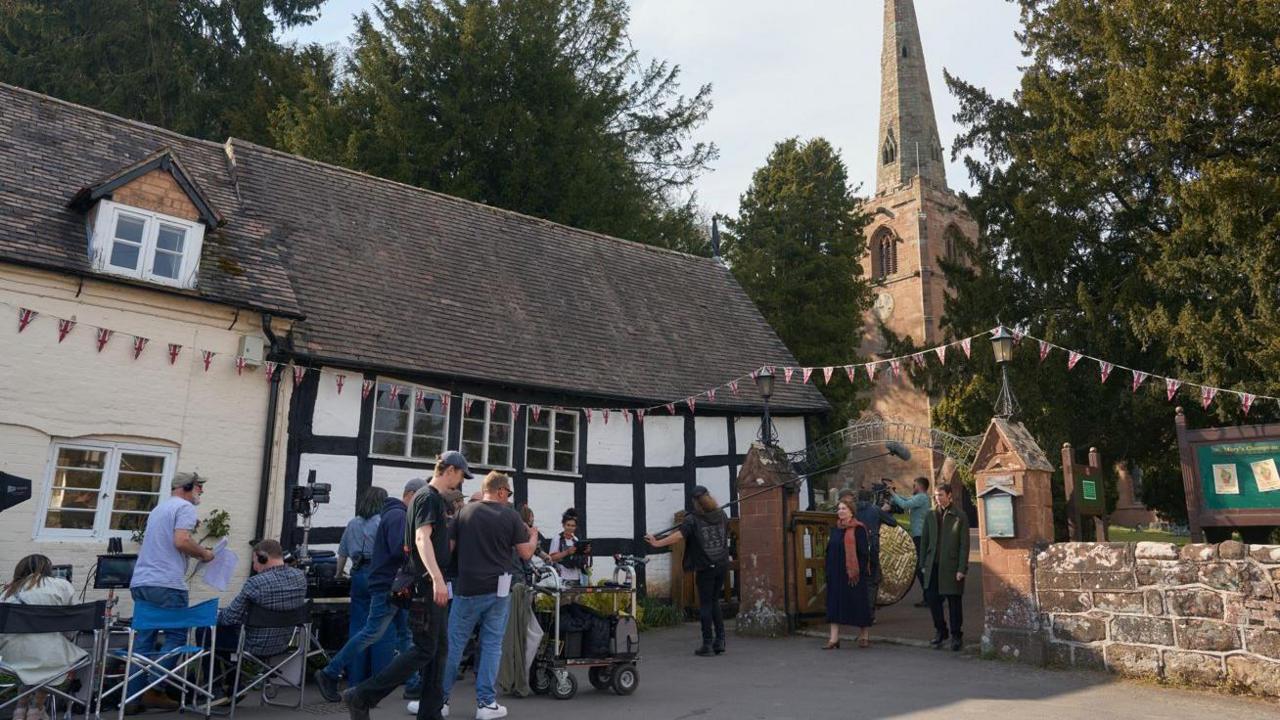 A number of people gathered around a black and white timber framed building with a church behind it, surrounded by trees