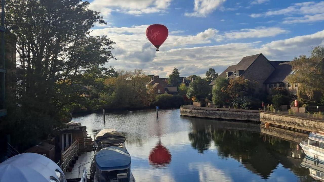 A red hot air balloon flies over the city if Oxford as seen from a bridge over the River Thames. The balloon's reflection can be seen in the water and some moored small boats are next to a jetty on the river bank's edge.
