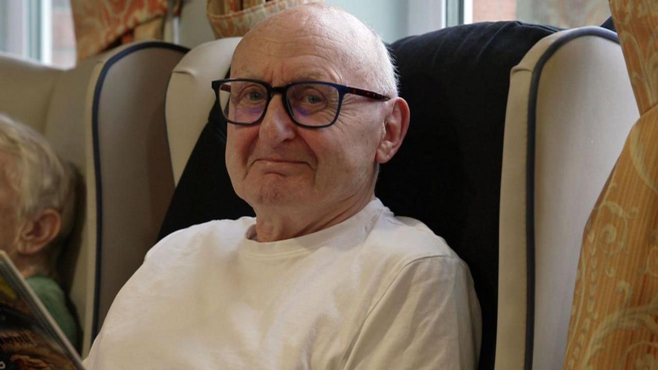 Michael Stevenson is bald, has black-framed glasses and is wearing a white T-shirt. He is sitting in a high-backed armchair which is cream in colour.