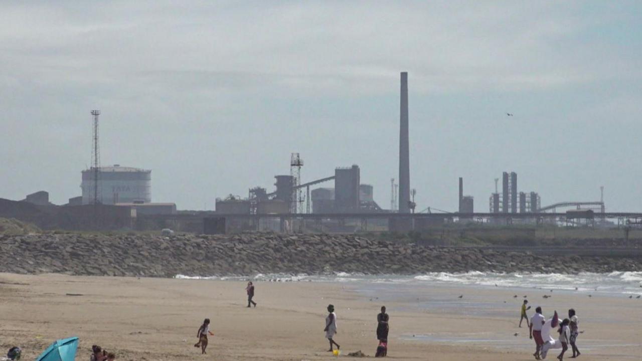 In the foreground there are people walking around on the beach and the sea is visible to the right of the picture. There's a rocky area in the background and beyond that there's the site of Port Talbot steelworks with a few chimneys and other large, industrial structures.