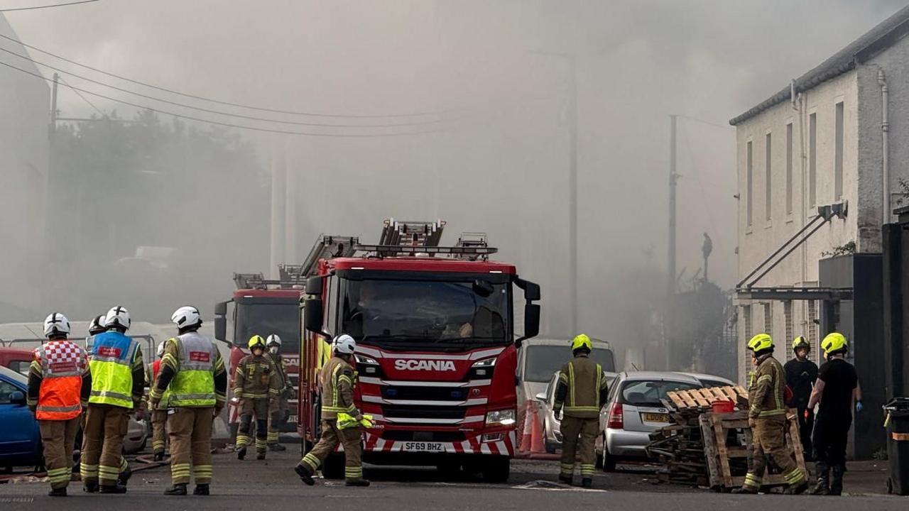Grey smoke surrounds a fire engines and firefighters on a street