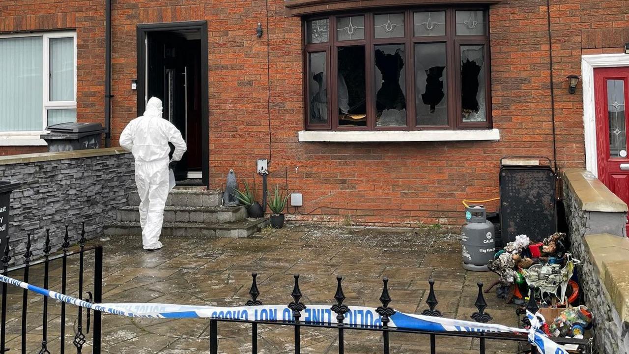 The front of a terraced house, with a front door on the left hand side and front windows to the right. The door is lying open and a forensic officer, with their back to the camera and wearing white overalls, is walking in. The windows are mostly smashed. In front of the house is a paved area leading to a black metal fence and gate, which is draped with blue and white police tape. 