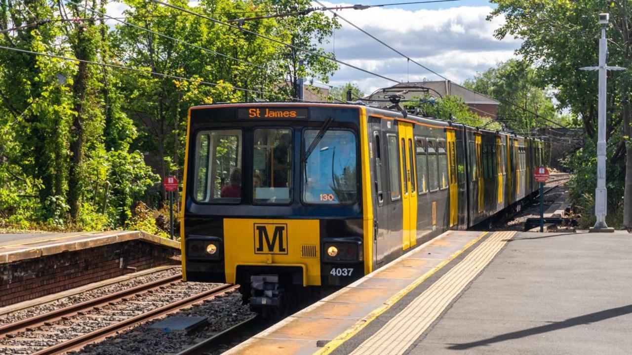 A  yellow and black Metro service train pulling into an outdoor station. It is heading to St James. Leafy trees surround the station.