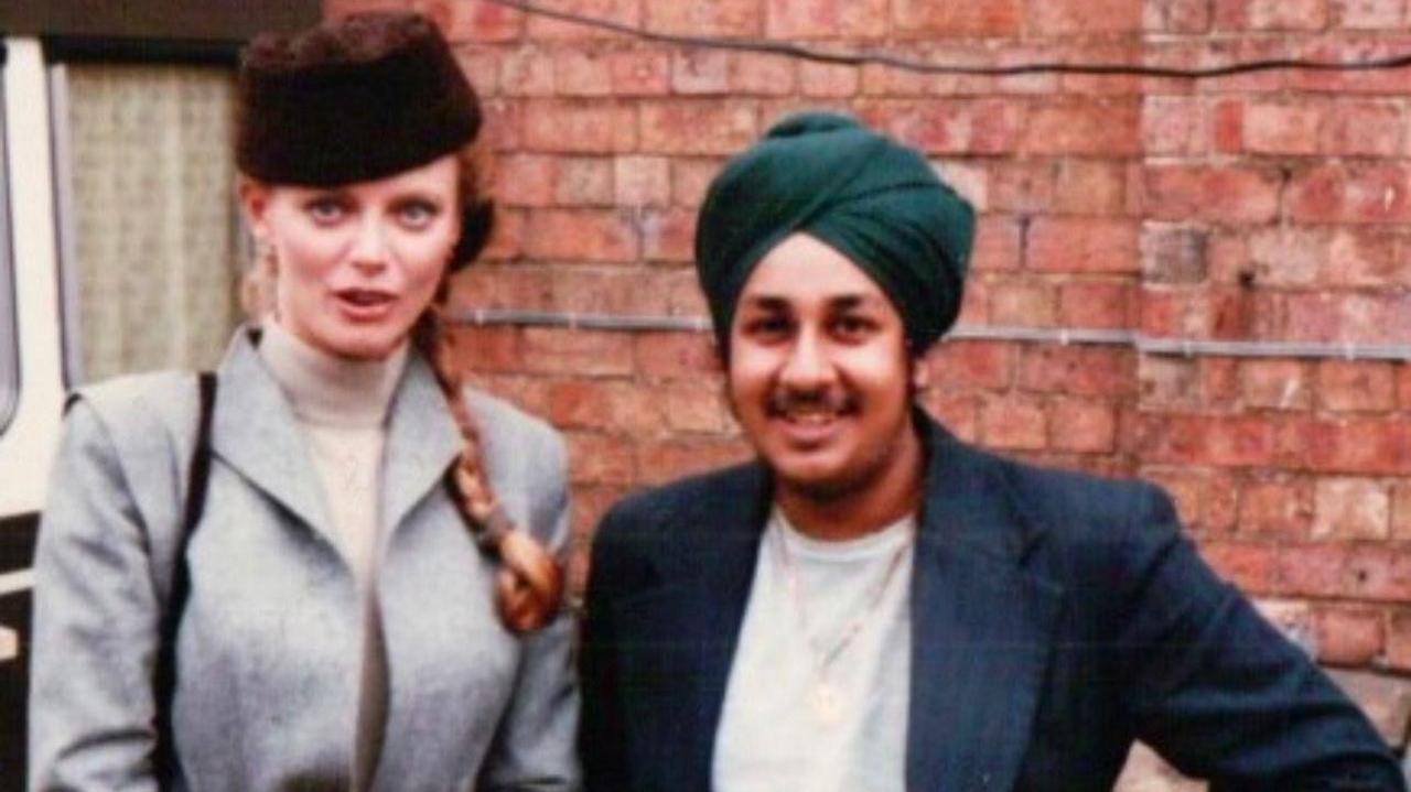 Kristina is wearing a grey coat and turtle neck cream jumper with a fluffy hat. Her blonde hair is in a plat over her shoulder. She is standing in front of a brick wall on the film set next to Del. Del is smiling at the camera with one hand on his hip. He is wearing a turquoise turban, gold necklace, grey top and blue and black striped blazer.