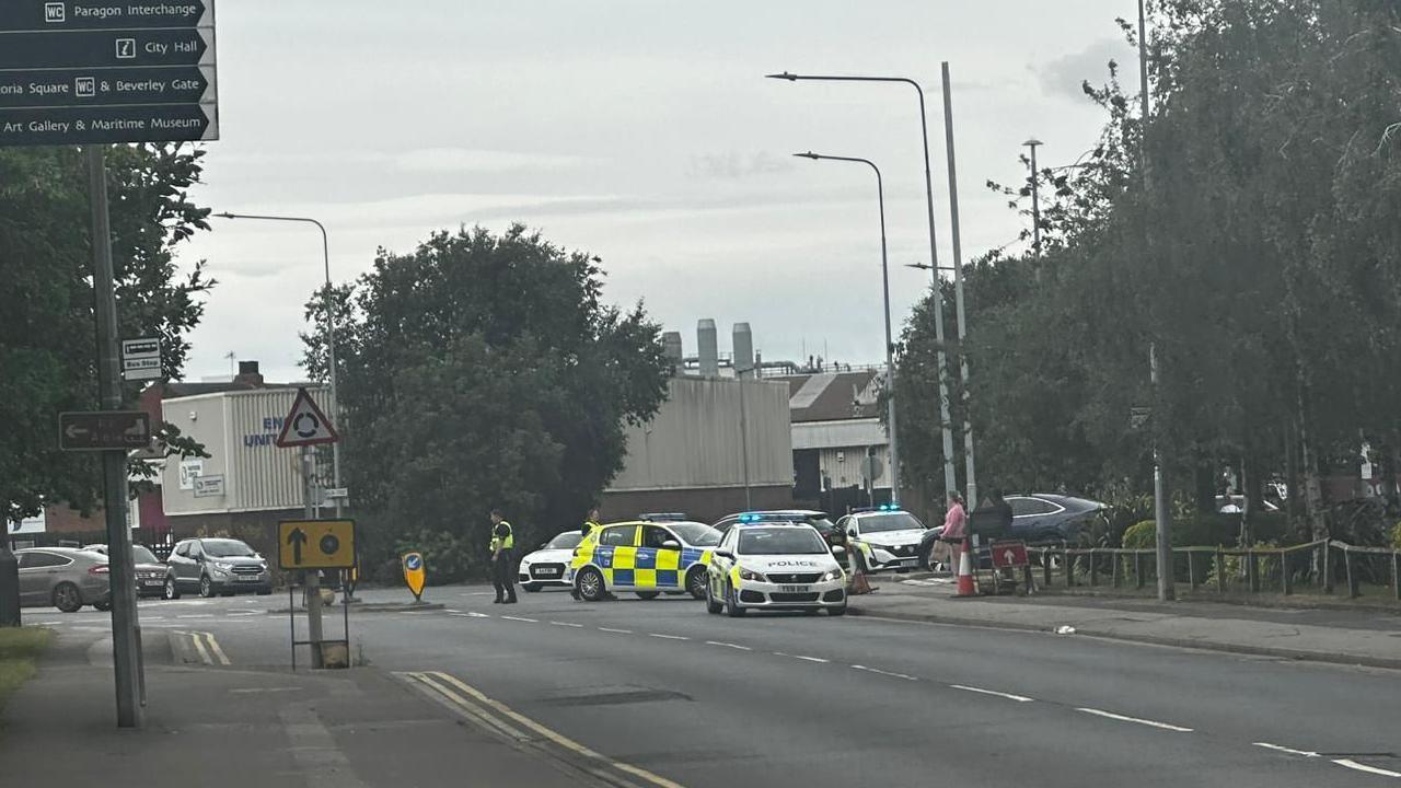Three police cars block the entrance to a car park. A number of industrial buildings are visible in the background. A row of trees is lining the road.